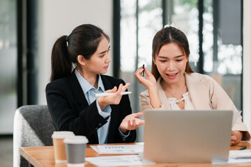 Two businesswomen discussing work at a desk with a laptop and coffee cups in a bright, modern office environment.