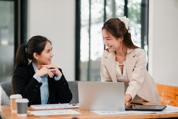 Two women engaged in a business meeting, discussing ideas in a modern office environment with laptops and documents.