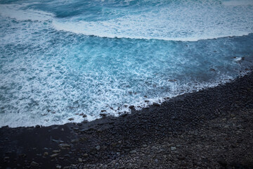 Stunning aerial view of ocean waves crashing on a dark pebble beach.  Perfect for travel, nature, or coastal themed projects. High-resolution image with vibrant colors.