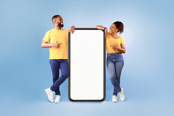 Hot offer, online sale. Funky African American young couple pointing at cellphone with mockup space over blue studio background. Guy and lady advertising new awesome mobile app or cool website