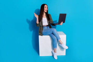 Trendy young woman using a laptop while sitting on a white block with a blue background