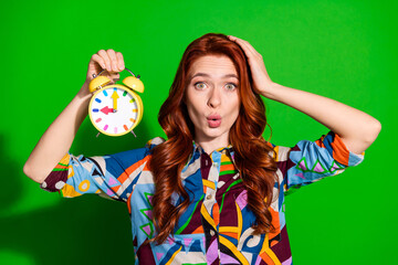 Young woman in colorful outfit holding alarm clock with puzzled expression against vibrant green background