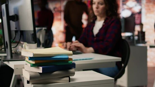 Focus on stack of books on startup office desk with woman in blurred background typing on PC, solving tasks. Pile of books in venture company workspace in front of employee working, camera A closeup