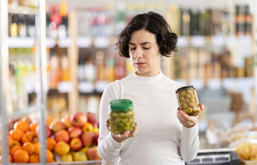 Woman choosing compare varieties pickled green olives in jar, doubt and think before buying. Client...
