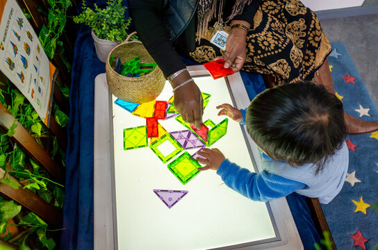 a child and adult or kindergarten educator playing with colorful magnetic geometric blocks on a light table, creating shapes and patterns as part of an educational activity or creative learning