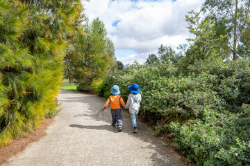 Two young children (3 to 4 years old) wearing wide brim sun hats walk hand in hand along a public...