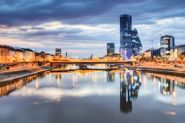 Naklejka premium Cityscape reflects beautifully in the tranquil river at twilight with dramatic clouds overhead.