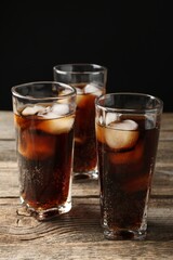 Refreshing cola with ice cubes in glasses on wooden table against black background, closeup