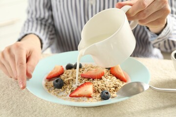 Woman pouring milk into bowl with oatmeal and berries at beige textured table, closeup