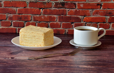 A piece of layered cake with protein cream on a saucer and a cup of hot coffee on a wooden table.