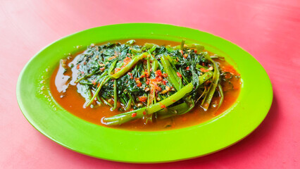 Stir-fried Water Spinach (cah or Tumis Kangkung) is one of Indonesian Daily Menu and Favorite Food. Served on green plate on red table. 45 degree set up