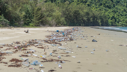 The trash is thrown by the sea on the beach. A lot of plastic bottles, boxes, and sticks are scattered on the white sand. Green vegetation. Turquoise ocean. Malaysia. Borneo. Kota Kinabalu.