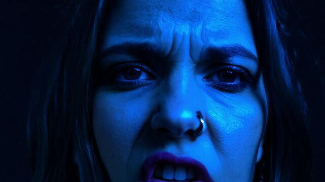 Static close-up video, cool blue lighting, woman, mismatched eyes, one blue, one brown, expressing fierce anger, plain black background, nostrils flare, lips curl, snarl, intense unwavering gaze
