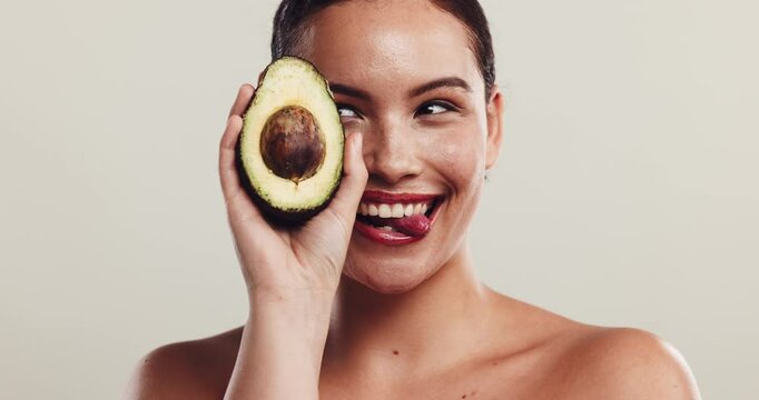 Woman, laugh and beauty with avocado in studio, funny face and cover eye for skincare by white background. Girl, person and fruit with nutrition, comic and tongue out with cosmetics in Costa Rica
