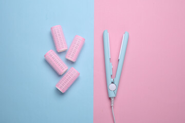Plastic curlers and hair straightener on blue pink background. Flat lay