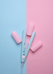 Plastic curlers and hair straightener on blue pink background. Flat lay