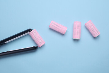 Plastic curlers and hair straightener on blue background. Flat lay