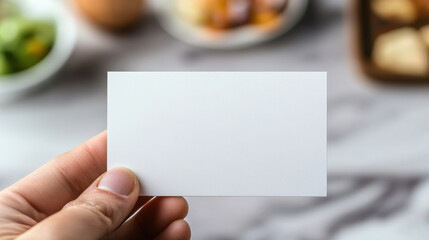 Obraz premium A close-up photograph of an empty white business card, held in a hand against a blurred background with some food items in the background