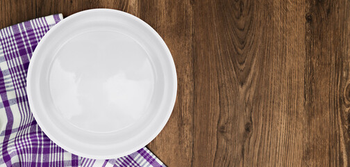 Empty white circle plate on wooden table with linen napkin. Overhead view. White ceramic plate on a rustic wooden table with a purple checkered napkin, ideal for food styling, dining concepts