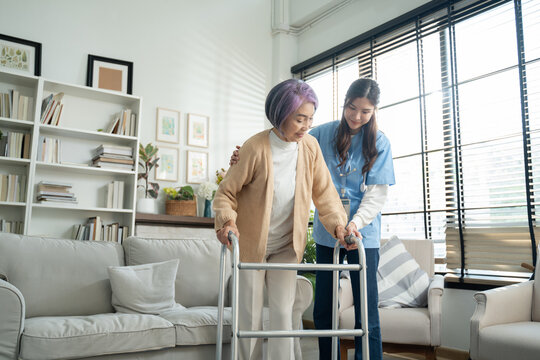 Asian young nurse supporting elderly patient disabled woman in using walker at home.Asian caregiver help care to senior female walk.Elderly patient care,love and care,disabled Physiotherapy concept