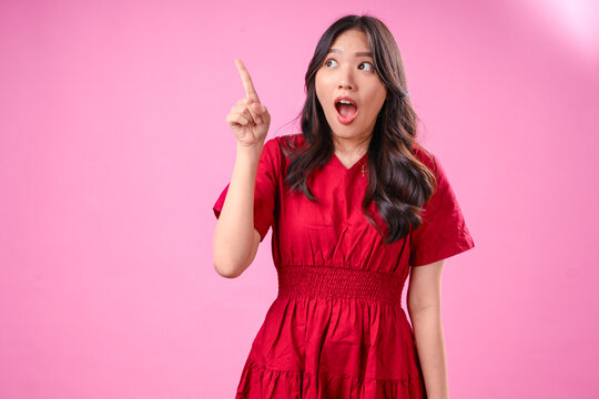 A young Asian woman with long wavy black hair wears a red dress, against a pink background. She looks up with wide eyes, and raises her index finger as if having a sudden realization