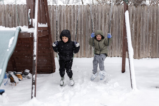 Two kids swing in the snow on a backyard play set with falling snow