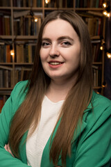 A professional woman wearing a green jacket poses with crossed arms against a backdrop of a well-stocked bookshelf and decorative string lights, suggesting a scholarly and inviting environment.