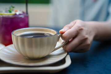 Close-up of woman holding ceramic cup with black coffee with vibrant purple cocktail in background. Woman relaxation, warm beverages, and cozy café experience in enjoy of sipping hot and cold drinks.