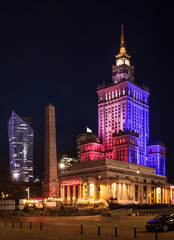 Night in Warsaw, Poland. The Palace of Culture and Science is illuminated in vibrant red, blue, and purple lights, contrasting with the dark sky. City lights and modern skyscrapers complete scene.
