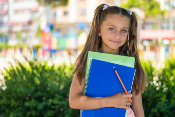 Smiling young girl with pigtails holding colorful notebooks and a pencil, standing outdoors. The setting suggests a return to school after summer vacation, radiating happiness and eagerness
