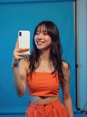 Happy young woman in a monochromatic orange outfit, playfully taking a mirror selfie with her phone, against a neon blue studio backdrop