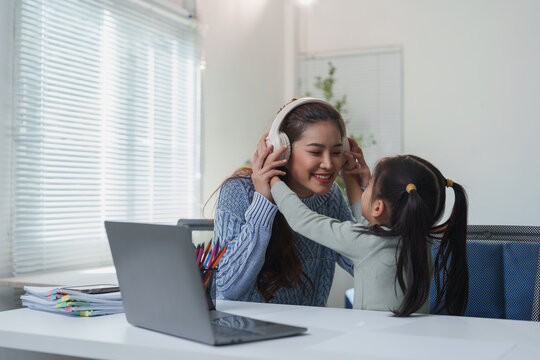 Smiling mother and daughter bonding over music at home. The mother wears headphones while the daughter playfully adjusts them. A laptop and colorful pencils are on the desk nearby - Powered by Adobe
