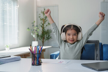 Smiling girl wearing headphones raises her arms in excitement while sitting at a desk with colored...