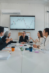 Diverse businesspeople are discussing stock market charts and graphs displayed on a large screen during a productive meeting in a modern office