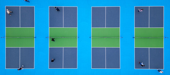 Aerial top-down view of multiple pickleball courts with players engaged in a match. The vibrant blue and green surfaces create a striking geometric pattern, capturing the dynamic movement and energy