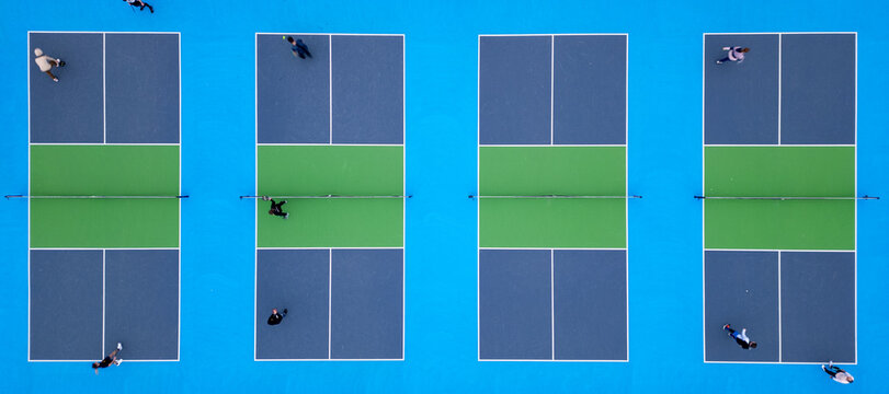 Aerial top-down view of multiple pickleball courts with players engaged in a match. The vibrant blue and green surfaces create a striking geometric pattern, capturing the dynamic movement and energy