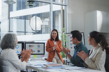 Diverse colleagues are clapping for a businesswoman after a successful presentation, celebrating achievement and teamwork in a modern office environment with charts displayed on computer screens