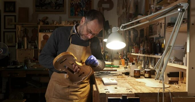 Close up of Luthier wearing an apron and gloves, carefully tampon ribs of a hand crafted violin in his workshop, illuminated by a lamp, surrounded by tools and materials, slow motion shot