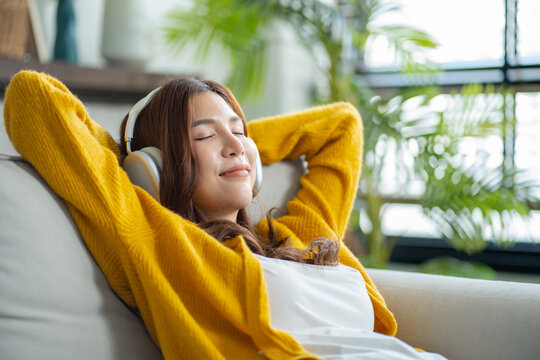 Happy young asian woman use headphones and relaxing at home.Female smile sitting on sofa,couch and holding mobile smartphone.Listening to music on lazy and radiating joy and relaxation.