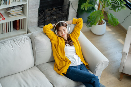 Happy young asian woman use headphones and relaxing at home.Female smile sitting on sofa,couch and holding mobile smartphone.Listening to music on lazy and radiating joy and relaxation.