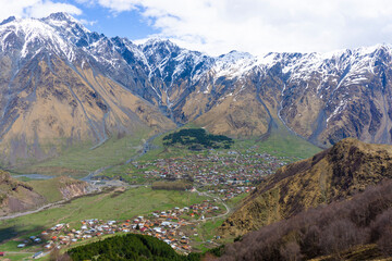 Naklejka premium View from above to the village of Gergeti and Stepantsminda. The valley of the Terek River is surrounded by high mountains covered with snow. Blue cloudy sky.