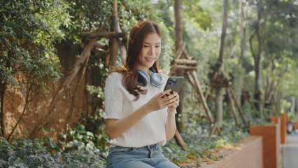 Naklejka premium Attractive young woman enjoys her smartphone while ing headphones in lush outdoor setting, radiating happiness and relaxation. Asian Woman Lifestyle