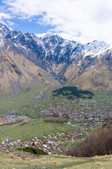 View from above to the village of Gergeti and Stepantsminda. The valley of the Terek River is surrounded by high mountains covered with snow. Blue cloudy sky.
