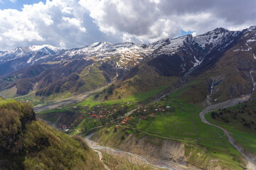 View from above of a small village on a cliff. The valley of the Terek River is surrounded by high mountains covered with snow. Blue cloudy sky.