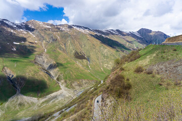 Mountain road leads up along the river gorge. Mountains are covered with green grass and partly with snow. Blue sky with clouds in the background