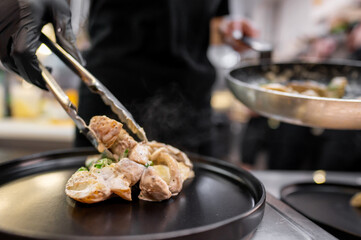 A chef in black gloves uses tongs to plate a gourmet dish in a professional kitchen. The dish features tender meat, garnished with herbs, creating an appetizing presentation.