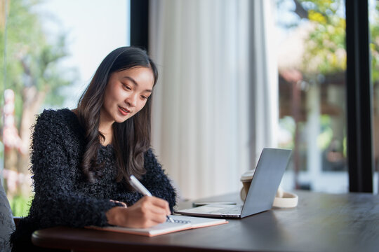 Young asian woman is taking notes in a notebook while studying online using her laptop at home, creating a productive and focused learning environment