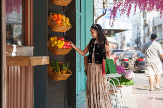 Stylishly dressed woman is reaching for some red apples in a wicker basket displayed on a street market stall, holding shopping bags in her other hand, enjoying a sunny day of shopping