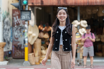 Young woman tourist smiling and walking in a street market in thailand. Wearing sunglasses on her head. A black vest and a beige skirt. With a blurred man consulting his smartphone in the background