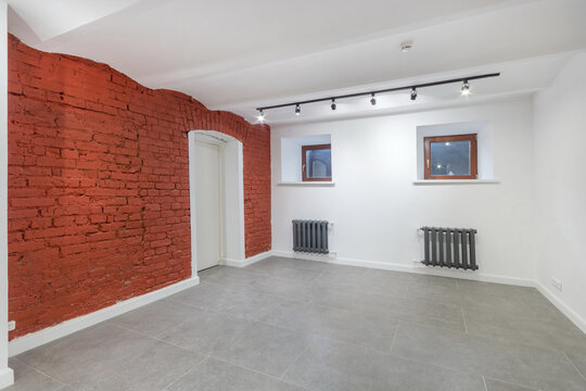 An empty room in the basement of the mansion. Gray floor tiles, a white door in a red brick wall. Unfurnished.